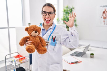 Téléchargez les photos : Jeune pédiatre hispanique tenant un ours en peluche à la clinique célébrant la victoire avec un sourire heureux et une expression gagnante avec les mains levées - en image libre de droit