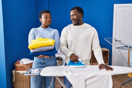 Photo for Young african american couple ironing clothes at laundry room smiling looking to the side and staring away thinking. - Royalty Free Image