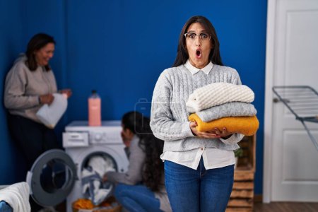 Foto de Tres mujeres lavando la ropa en casa asustadas y conmocionadas por la sorpresa y la expresión asombrada, el miedo y la cara emocionada. - Imagen libre de derechos