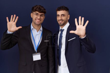 Foto de Dos hombres de negocios hispanos con ropa de negocios mostrando y señalando con los dedos el número diez mientras sonríen confiados y felices. - Imagen libre de derechos