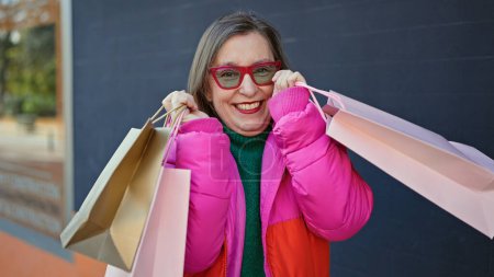 Photo for Mature hispanic woman with grey hair smiling going shopping holding bags at street - Royalty Free Image