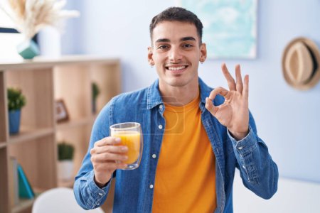 Téléchargez les photos : Bel homme hispanique boire un verre de jus d'orange faire ok signe avec les doigts, sourire geste amical excellent symbole - en image libre de droit