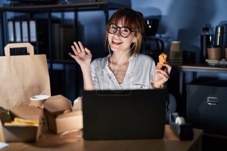 Foto de Joven hermosa mujer que trabaja con computadora portátil y comer comida entrega renuncia decir hola feliz y sonriente, gesto de bienvenida amistoso - Imagen libre de derechos