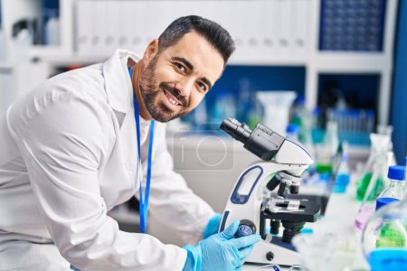 Téléchargez les photos : Jeune homme hispanique scientifique souriant confiant en utilisant le microscope au laboratoire - en image libre de droit