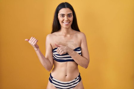 Photo for Young brunette woman wearing bikini over yellow background pointing to the back behind with hand and thumbs up, smiling confident - Royalty Free Image
