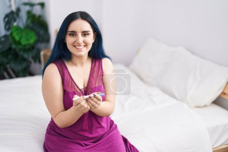 Foto de Joven mujer caucásica sonriendo confiado sosteniendo prueba de embarazo en el dormitorio - Imagen libre de derechos