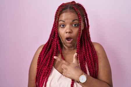 Photo for African american woman with braided hair standing over pink background surprised pointing with finger to the side, open mouth amazed expression. - Royalty Free Image