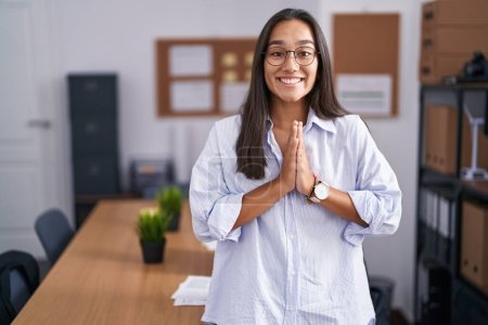 Photo for Young hispanic woman at the office praying with hands together asking for forgiveness smiling confident. - Royalty Free Image