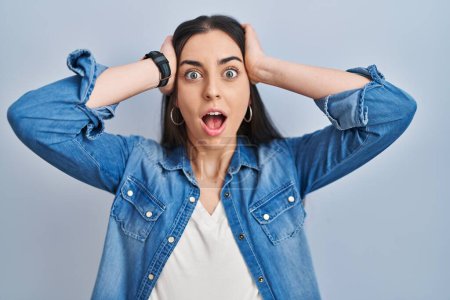 Photo for Hispanic woman standing over blue background crazy and scared with hands on head, afraid and surprised of shock with open mouth - Royalty Free Image