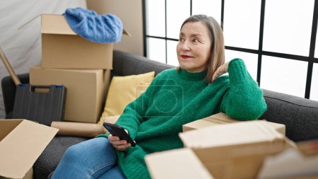 Foto de Mujer hispana madura con cabello gris usando un smartphone sentado en un sofá en un nuevo hogar - Imagen libre de derechos
