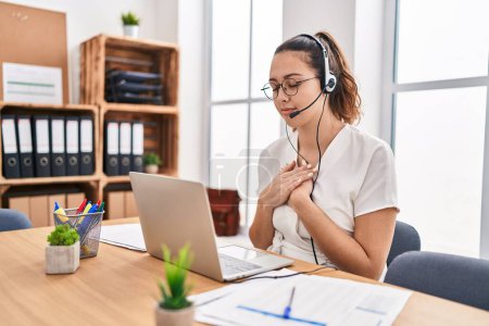 Photo for Young hispanic woman wearing call center agent headset at the office smiling with hands on chest with closed eyes and grateful gesture on face. health concept. - Royalty Free Image