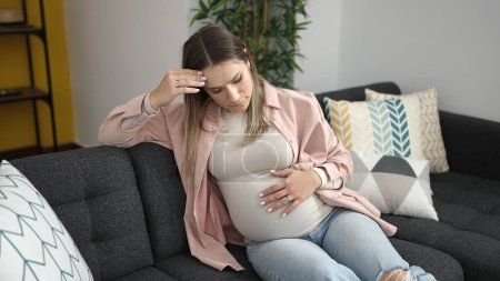 Foto de Mujer embarazada joven tocando el vientre con expresión preocupada en casa - Imagen libre de derechos