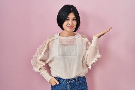 Téléchargez les photos : Jeune femme asiatique debout sur fond rose souriant gai présentation et pointant avec la paume de la main en regardant la caméra. - en image libre de droit