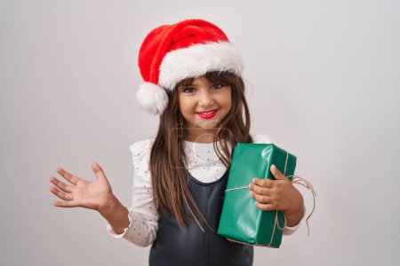 Foto de Niña hispana con sombrero de navidad y regalos celebrando el logro con sonrisa feliz y expresión ganadora con la mano levantada - Imagen libre de derechos
