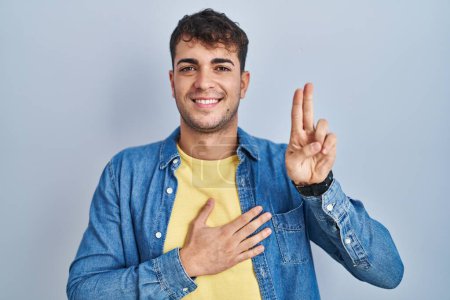 Foto de Joven hombre hispano de pie sobre fondo azul sonriendo jurando con la mano en el pecho y los dedos hacia arriba, haciendo una promesa de lealtad juramento - Imagen libre de derechos