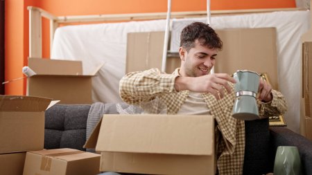 Foto de Joven hombre hispano sonriendo confiado desempacando caja de cartón en nuevo hogar - Imagen libre de derechos