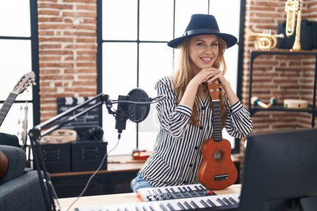 Foto de Joven mujer rubia músico sonriendo confiado apoyándose en ukelele en el estudio de música - Imagen libre de derechos