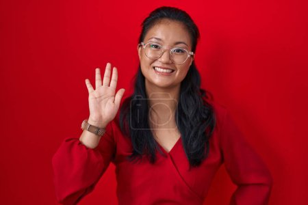 Foto de Asiática joven mujer de pie sobre rojo fondo renunciando a decir hola feliz y sonriente, amable gesto de bienvenida - Imagen libre de derechos