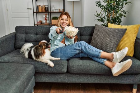 Photo for Young caucasian woman watching movie sitting on sofa with dog at home - Royalty Free Image