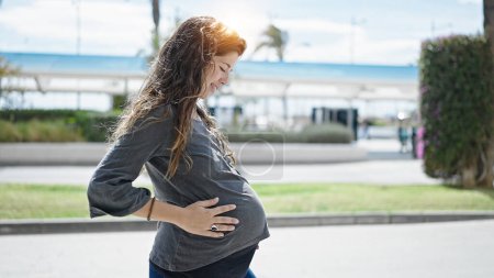 Foto de Joven embarazada sonriendo confiado tocando vientre en el parque - Imagen libre de derechos