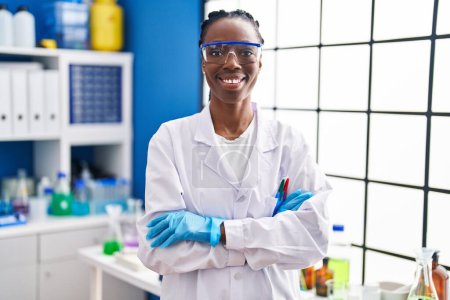 Téléchargez les photos : Femme scientifique afro-américaine souriante confiante debout avec les bras croisés geste au laboratoire - en image libre de droit