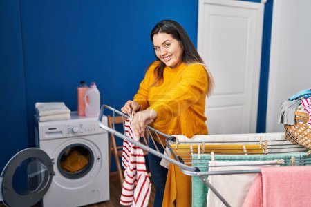 Young beautiful plus size woman smiling confident hanging clothes on clothesline at laundry room