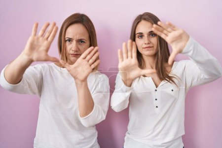 Photo for Middle age mother and young daughter standing over pink background doing frame using hands palms and fingers, camera perspective - Royalty Free Image