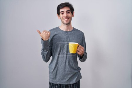 Téléchargez les photos : Jeune homme hispanique portant un pyjama buvant une tasse de café pointant vers le dos avec la main et les pouces levés, souriant confiant - en image libre de droit