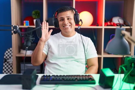 Téléchargez les photos : Jeune homme hispanique jouant à des jeux vidéo renonçant à dire bonjour heureux et souriant, geste de bienvenue amical - en image libre de droit