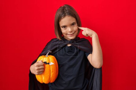 Téléchargez les photos : Petit garçon hispanique portant une cape et tenant citrouille halloween sourire heureux pointant avec la main et le doigt - en image libre de droit