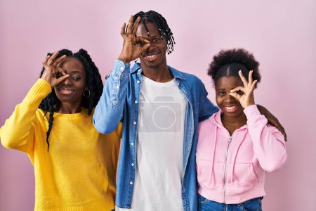 Foto de Grupo de tres jóvenes negros de pie juntos sobre fondo rosa haciendo buen gesto con la mano sonriente, los ojos mirando a través de los dedos con la cara feliz. - Imagen libre de derechos
