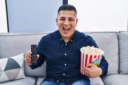 Foto de Joven hispano comiendo palomitas de maíz usando control de televisión sonriendo y riendo en voz alta porque gracioso chiste loco. - Imagen libre de derechos