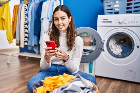 Téléchargez les photos : Young beautiful hispanic woman smiling confident using smartphone at laundry room - en image libre de droit