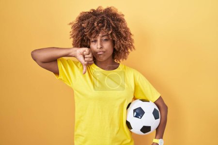 Photo for Young hispanic woman with curly hair holding football ball with angry face, negative sign showing dislike with thumbs down, rejection concept - Royalty Free Image