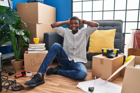 Téléchargez les photos : Afro-Américain assis sur le sol à la nouvelle maison relaxant et étirant, bras et mains derrière la tête et le cou souriant heureux - en image libre de droit
