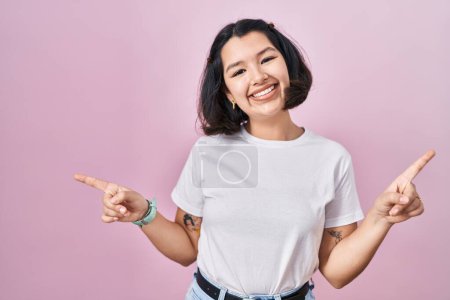 Téléchargez les photos : Jeune femme hispanique portant t-shirt blanc décontracté sur fond rose souriant confiant pointant avec les doigts vers différentes directions. espace de copie pour la publicité - en image libre de droit