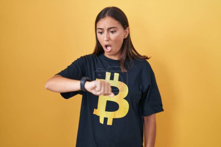 Foto de Mujer hispana joven vistiendo camiseta bitcoin mirando el tiempo reloj preocupado, miedo de llegar tarde - Imagen libre de derechos