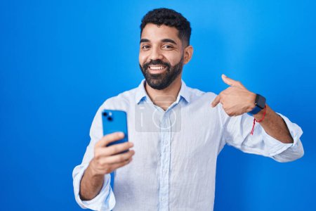 Foto de Hombre hispano con barba usando mensaje de escritura de teléfonos inteligentes que se ve confiado con sonrisa en la cara, señalándose con los dedos orgullosos y felices. - Imagen libre de derechos
