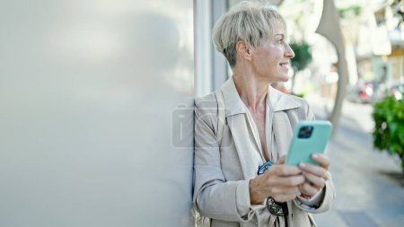 Foto de Mujer rubia de mediana edad sonriendo confiada usando teléfono inteligente en la calle - Imagen libre de derechos