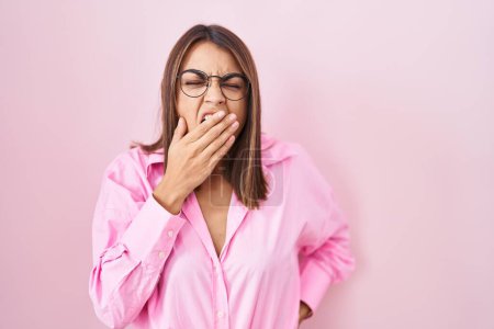 Foto de Mujer hispana joven con gafas de pie sobre fondo rosa aburrido bostezo cansado cubriendo la boca con la mano. inquietud y somnolencia. - Imagen libre de derechos