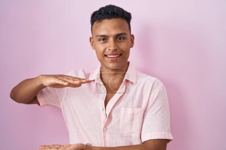 Téléchargez les photos : Jeune homme hispanique debout sur fond rose geste avec les mains montrant grand et grand signe de taille, symbole de mesure. souriant en regardant la caméra. concept de mesure. - en image libre de droit