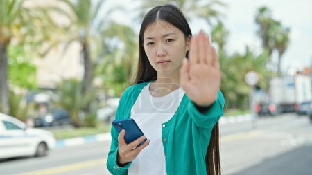 Foto de Joven mujer china haciendo gesto de parada con mano usin teléfono inteligente en la calle - Imagen libre de derechos