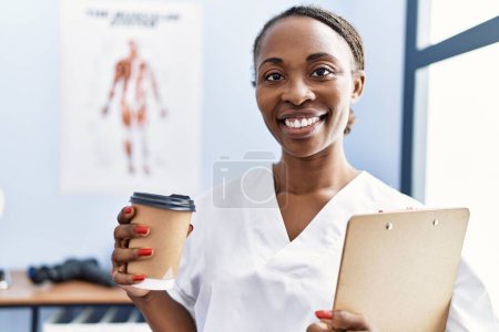 Photo for African american woman physiotherapist holding medical report drinking coffee at rehab clinic - Royalty Free Image
