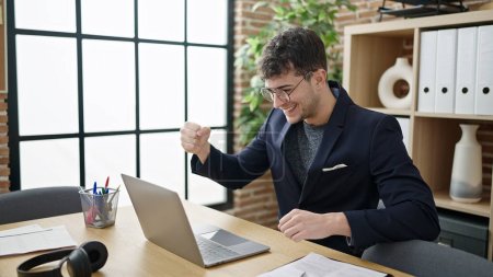Young hispanic man business worker using laptop celebrating at office
