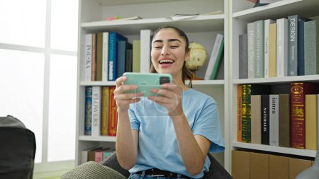 Téléchargez les photos : Jeune belle étudiante hispanique regardant une vidéo sur smartphone à la bibliothèque de l'université - en image libre de droit