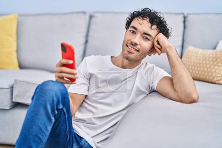 Foto de Hombre hispano joven usando teléfono inteligente sentado en el suelo en casa - Imagen libre de derechos