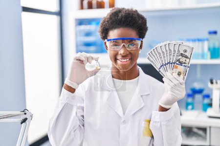 Foto de Mujer afroamericana que trabaja en el laboratorio científico sosteniendo dólares sonriendo con una sonrisa alegre y fresca en la cara. mostrando dientes. - Imagen libre de derechos