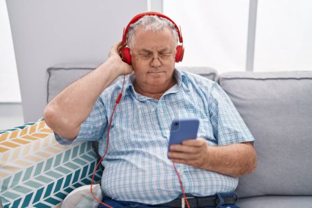 Foto de Hombre de pelo gris de mediana edad escuchando música sentado en el sofá en casa - Imagen libre de derechos