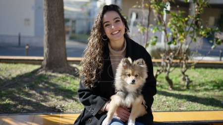 Téléchargez les photos : Jeune femme hispanique avec chien souriant confiant assis sur le banc au parc - en image libre de droit