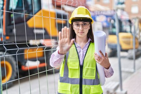 Photo for Hispanic girl wearing architect hardhat at construction site with open hand doing stop sign with serious and confident expression, defense gesture - Royalty Free Image